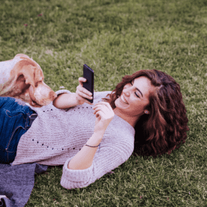 Lady lying on the grass taking a photo of a dog that is on her stomach