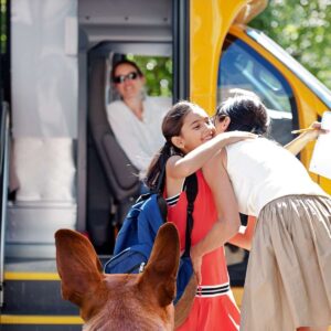 girl in red dress hugging mom before getting on school bus with a dog watching on