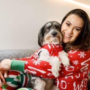 dark haired women in holiday pajamas holding her dog dressed in holiday pajamas too for a thanksgiving tradition