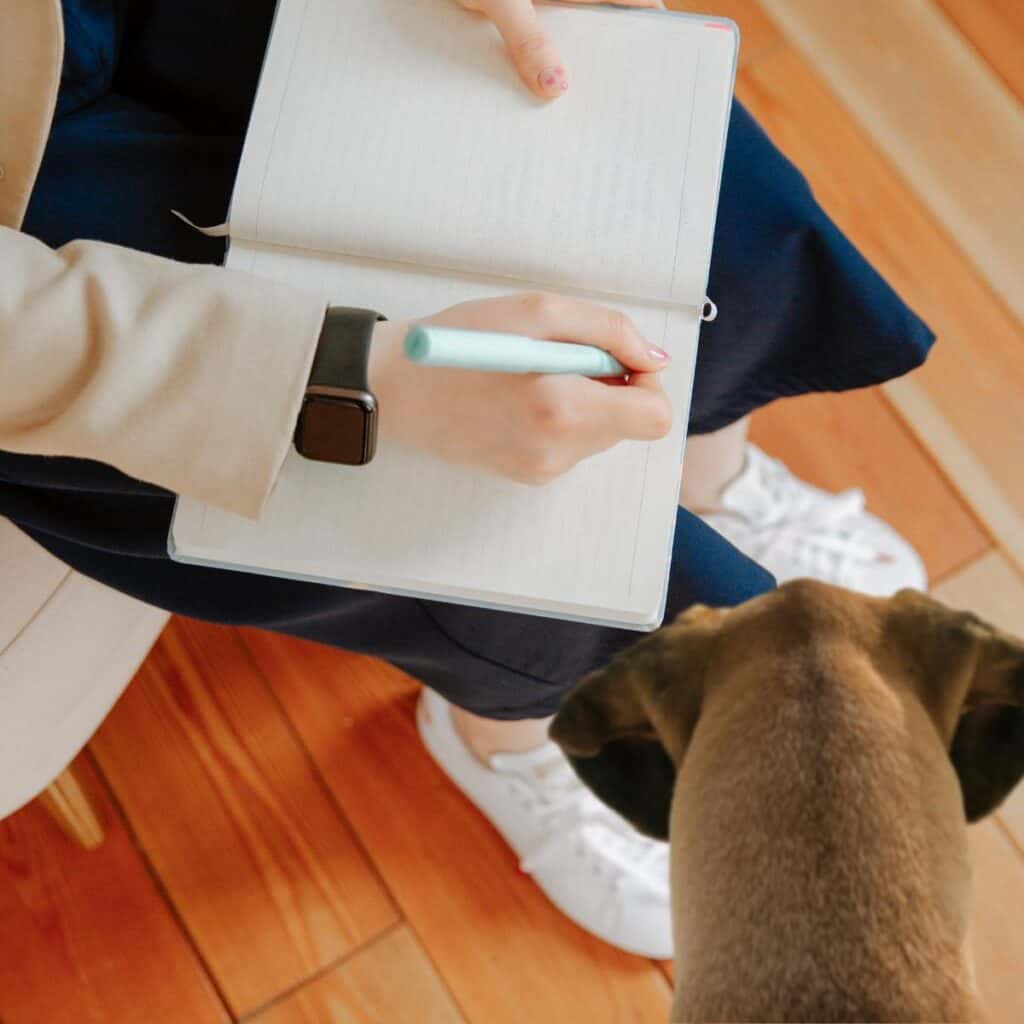 Person with a note pad, apple watch and wearing blue pants sitting down and taking notes about a brown dog looking on.