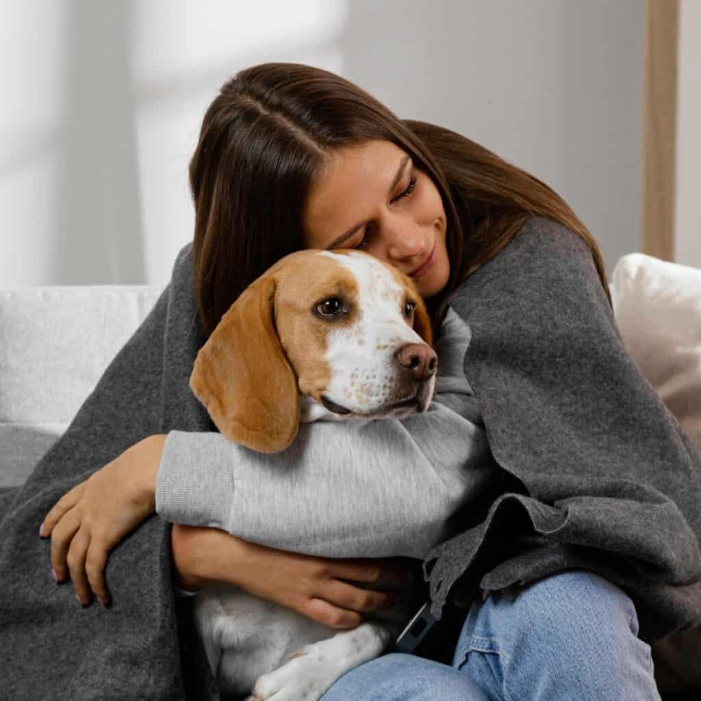 Brown haired woman wrapped in a grey blanket sitting on a couch with a brown and white dog.