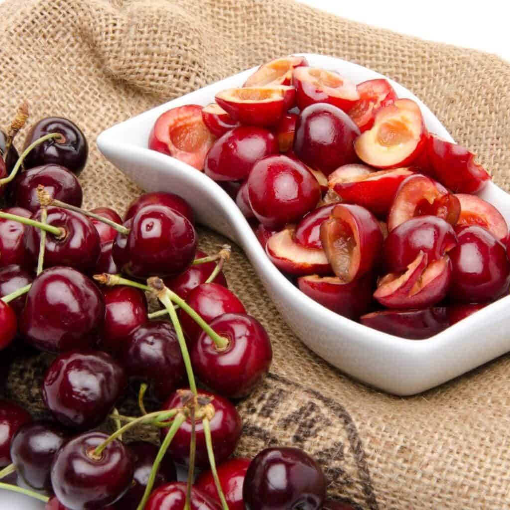 A pile of cherries whole with stem and a serving tray of cherries with pits and stem removed.